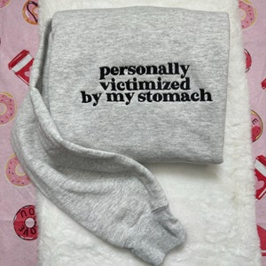 Puede incluir: Sudadera gris con texto bordado en negro que dice "personalmente victimizado por mi estómago".