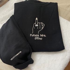 May include: Black sweatshirt with a white embroidered design of a hand giving the middle finger with a ring on the ring finger. The text "Future Mrs. Paris" is embroidered below the hand.
