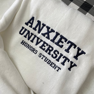 White sweatshirt with Anxiety University Honors Student embroidered in black thread.