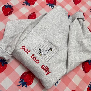 May include: A light grey sweatshirt with the embroidered phrase "got too silly" in red. The sweatshirt features a square patch with a duck design inside a cage. The background is a pink and white checkered pattern with strawberry illustrations.