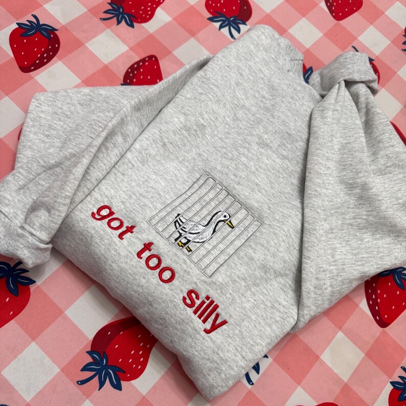 May include: A light grey sweatshirt with the embroidered phrase "got too silly" in red. The sweatshirt features a square patch with a duck design inside a cage. The background is a pink and white checkered pattern with strawberry illustrations.