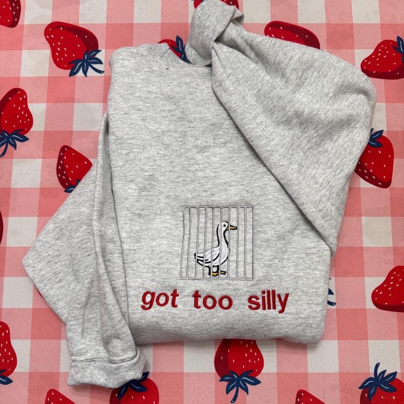May include: A light grey hooded sweatshirt with an embroidered duck design behind bars. The red text "got too silly" is embroidered below. The sweatshirt is folded on a pink and white checkered surface decorated with strawberry illustrations.