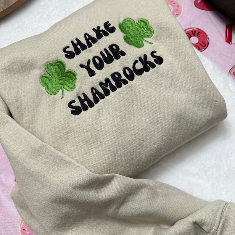 May include: A beige sweatshirt with the text "Shake Your Shamrocks" embroidered in black with green shamrock designs.