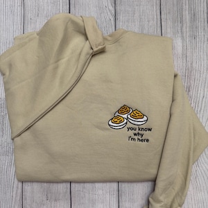 Könnte beinhalten: Beiges Sweatshirt mit gesticktem Design von gefüllten Eiern und dem Text "you know why I'm here". Das Kleidungsstück ist auf einer Holzoberfläche gefaltet.