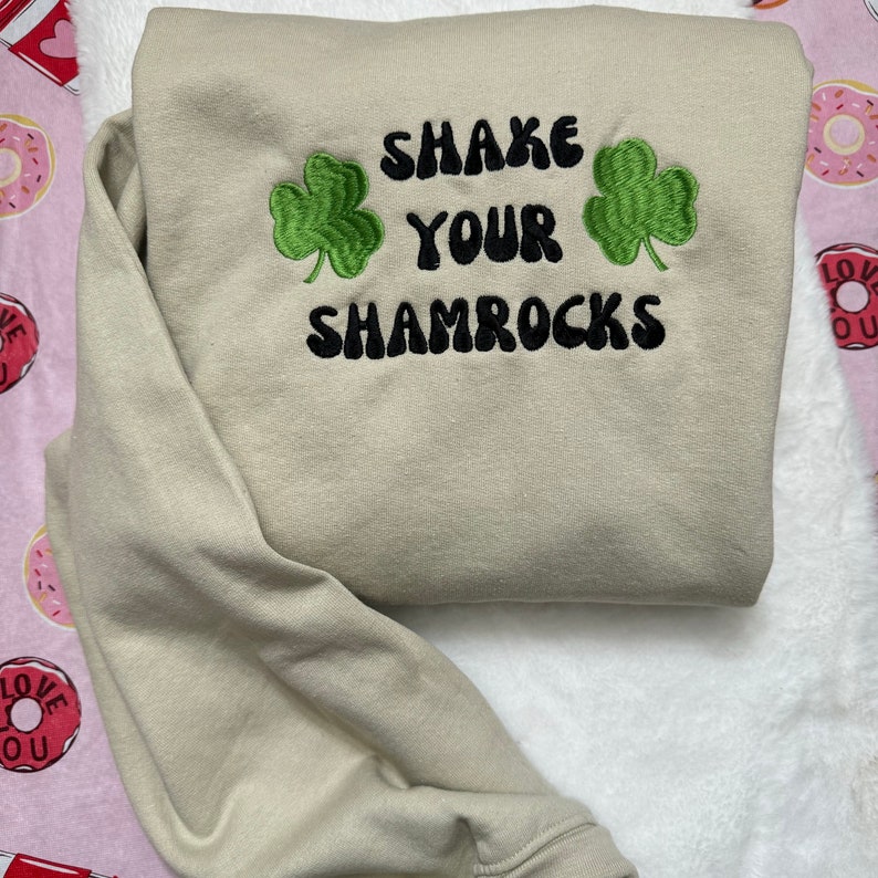 May include: A light beige sweatshirt with the text "Shake Your Shamrocks" in black with two green shamrock designs.