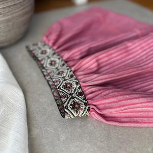 May include: Close-up of a pink striped fabric with a decorative band. The band features a geometric pattern in shades of green, brown, and white. The fabric appears to be part of a garment, possibly a sleeve or cuff.