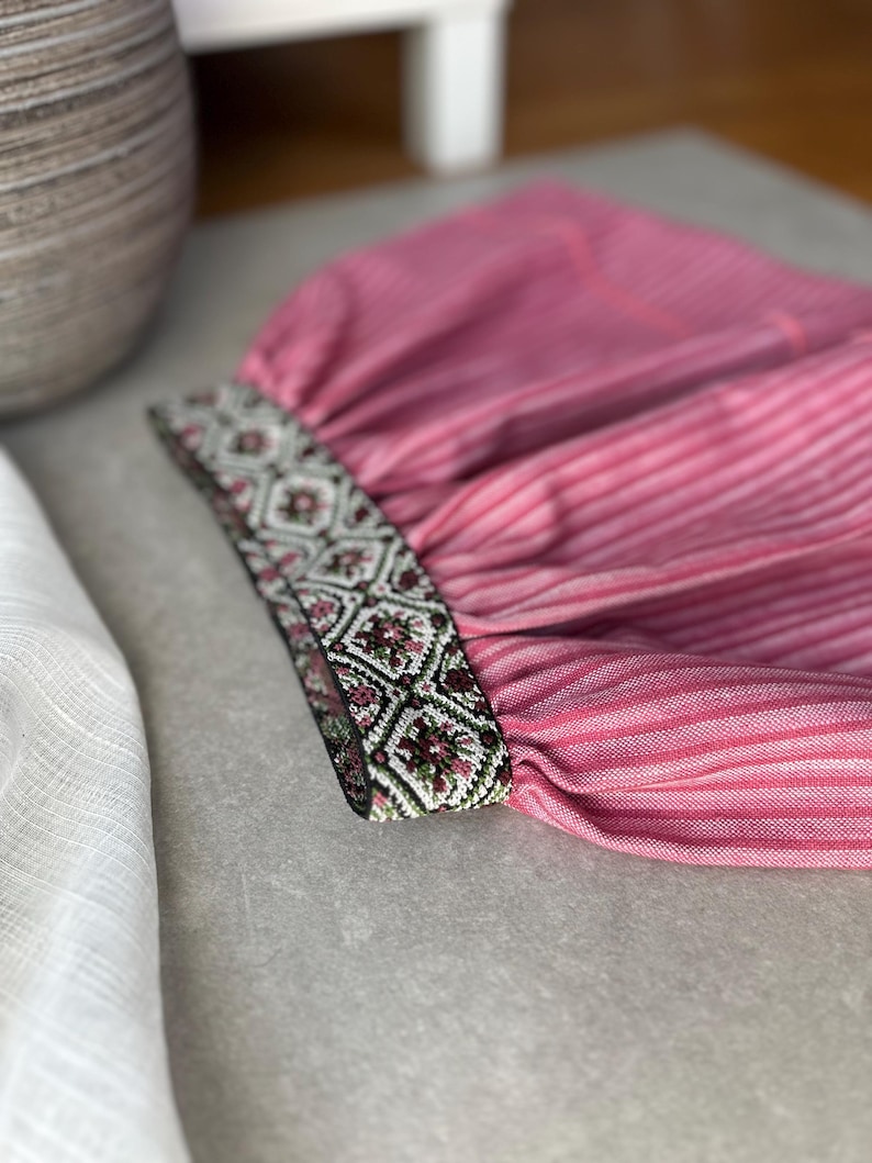 May include: Close-up of a pink striped fabric with a decorative band. The band features a geometric pattern in shades of green, brown, and white. The fabric appears to be part of a garment, possibly a sleeve or cuff.