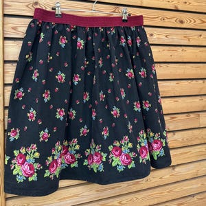 May include: A black skirt with a floral pattern of pink roses, blue accents, and green leaves. The skirt has a burgundy waistband and is hanging on a metal hanger against a wooden background.
