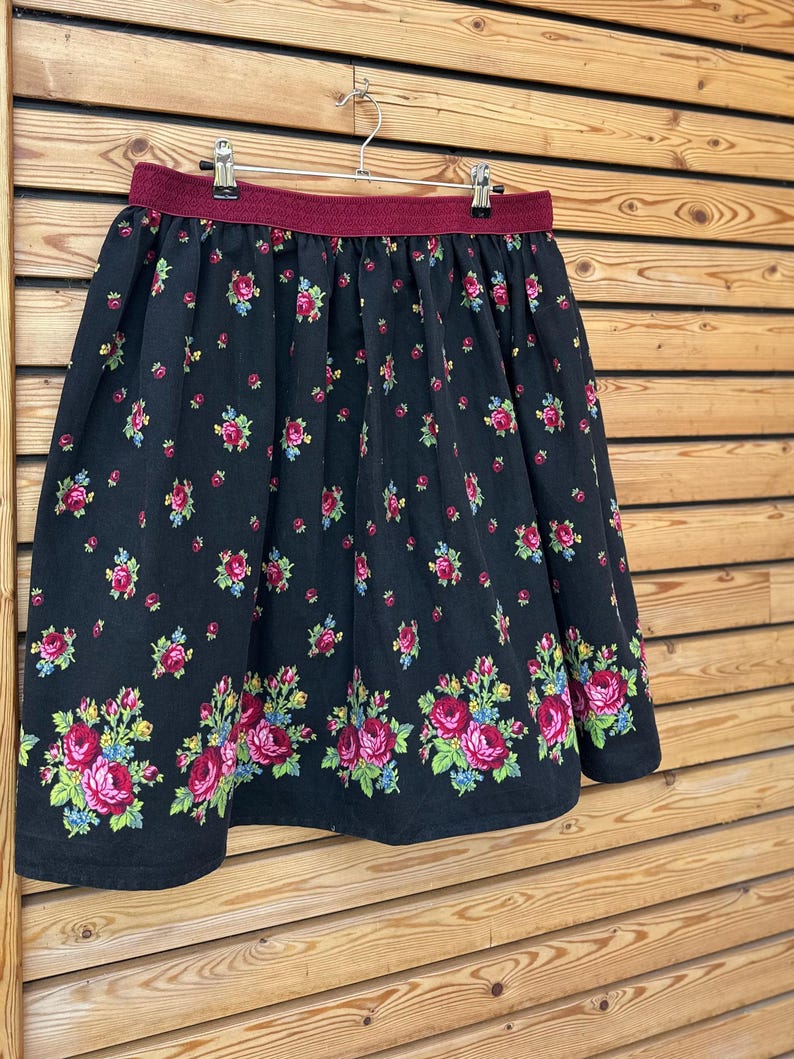 May include: A black skirt with a floral pattern of pink roses, blue accents, and green leaves. The skirt has a burgundy waistband and is hanging on a metal hanger against a wooden background.