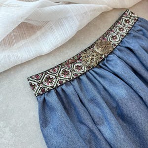 May include: A blue skirt with a decorative embroidered waistband featuring a floral and geometric pattern in shades of pink, green, and white. A silver-colored metal embellishment is centered on the waistband. The skirt is made of a soft, flowing fabric.