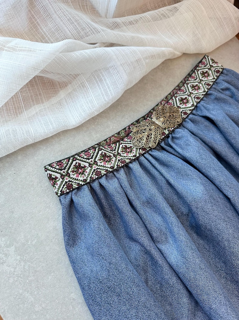 May include: A blue skirt with a decorative embroidered waistband featuring a floral and geometric pattern in shades of pink, green, and white. A silver-colored metal embellishment is centered on the waistband. The skirt is made of a soft, flowing fabric.