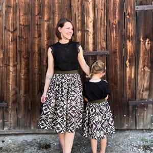 May include: A woman and a child wearing matching outfits. The woman wears a black lace top and a patterned skirt with a brown and white geometric design. The child wears a black top and a matching skirt. They stand in front of a wooden door.