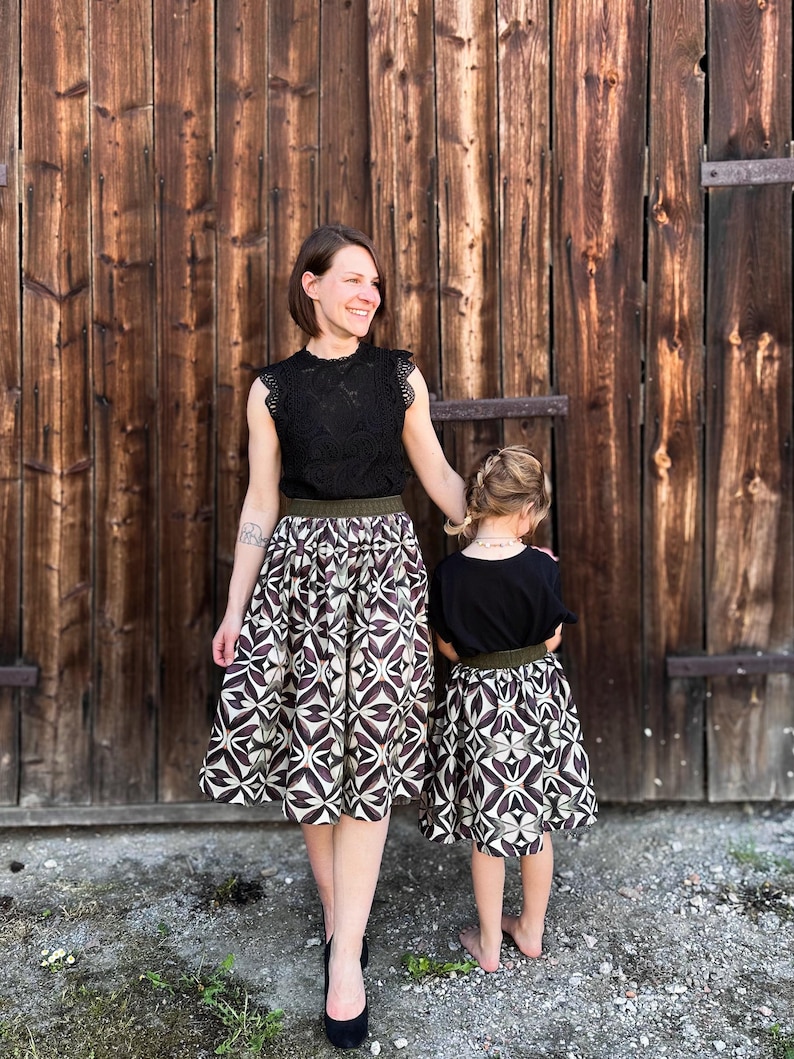 May include: A woman and a child wearing matching outfits. The woman wears a black lace top and a patterned skirt with a brown and white geometric design. The child wears a black top and a matching skirt. They stand in front of a wooden door.