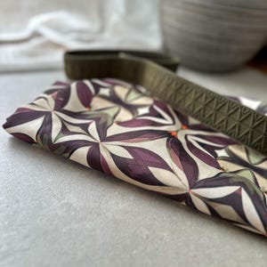 May include: Folded fabric with a floral pattern in shades of purple, cream, and green. A green strap with a diamond pattern is partially visible. The fabric is on a light gray surface.