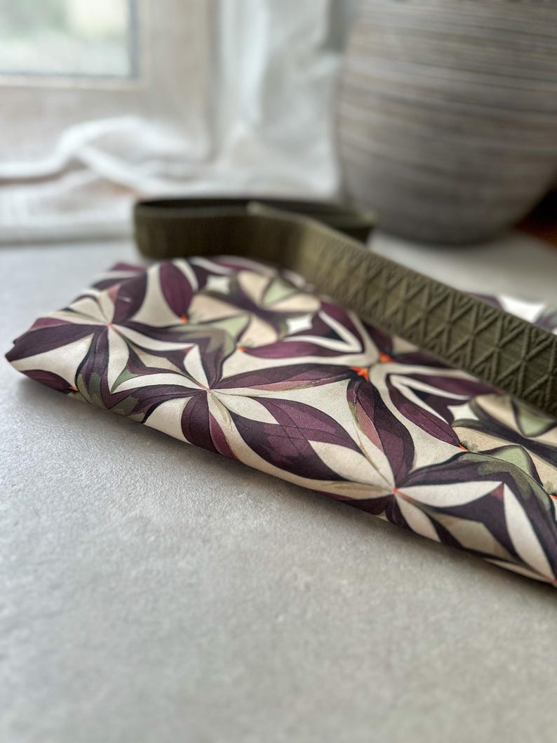 May include: Folded fabric with a floral pattern in shades of purple, cream, and green. A green strap with a diamond pattern is partially visible. The fabric is on a light gray surface.