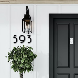 May include: Black house number plaque with the number 593 on a white wall with a black door and a black lantern light fixture.