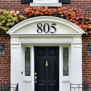 May include: A white door with a black door and a black house number 805 above the door. The house is made of red brick and has a white trim around the door and windows. There are vines growing on the side of the house.