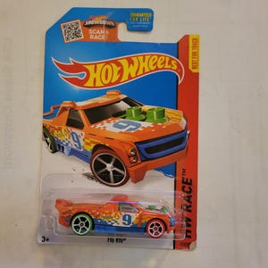 May include: A Hot Wheels toy car in orange, blue, and green with a pixelated design. The car is labeled "Fig Rig" and has the number "9" on the side. The car is on a white background with a blue and red Hot Wheels logo.