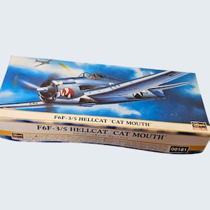 May include: A model kit box featuring a blue and silver F6F-3/5 Hellcat Cat Mouth airplane illustration. The box has the text "F6F-3/5 HELLCAT 'CAT MOUTH'" in white. The box also has the number "00181".