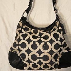 May include: Black and cream-colored shoulder bag with a repeating interlocking C pattern. The bag has black leather accents on the bottom and strap, with silver-tone hardware. A small Coach tag hangs from the strap.