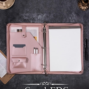 May include: A pink leather binder with a zipper closure, multiple pockets, and a ring binder mechanism. The binder is open to reveal a blank white sheet of paper and a pen.