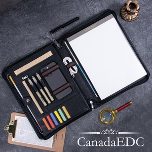 以下が含まれることがあります： ジッパー付きの黒い革製ポートフォリオ。ポートフォリオが開いており、ノートパッド、ペン、電話、充電ケーブルが見えます。画像の下部に「Canada EDC」というテキストが印刷されています。