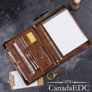 May include: A brown leather portfolio with a zipper closure, multiple pockets, and a notepad. The portfolio is open and the notepad is visible. The text "George Clooney - I'll sick" is visible on the inside of the portfolio.