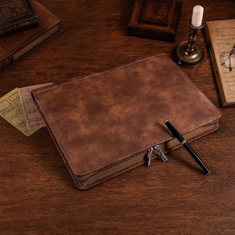 May include: A rectangular brown leather-like portfolio case with a smooth, mottled texture and a zippered closure. A sleek black fountain pen with gold-tone accents rests diagonally across the top surface of the closed case.