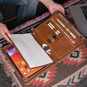 May include: A tan leather portfolio case, open to show an iPad, pens, and other accessories. The case has multiple compartments and a zip closure. The interior includes a white paper cover. The case is on a patterned rug.