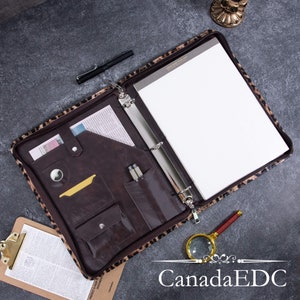 May include: A brown leather binder with a leopard print cover. The binder is open and has several pockets and slots for holding cards, pens, and other items. The binder also has a zipper closure.