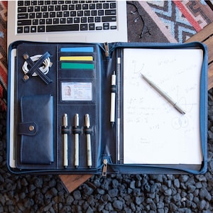 Op de afbeelding: Open marineblauw leren portfolio met ritssluiting, met een notitieblok met vergelijkingen, pennen en kaartsleuven. Op de achtergrond is een laptop zichtbaar. De portfolio bevat ook een telefoon, ID en oplaadkabels.