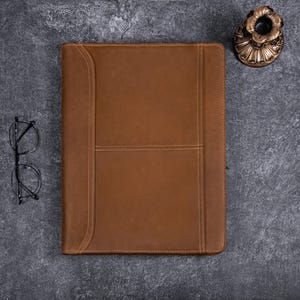 Puede incluir: Un portafolio de cuero marrón con un diseño cosido. El portafolio está cerrado y acostado sobre una superficie gris texturizada.