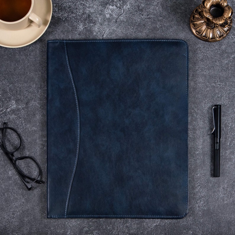 May include: A rectangular dark blue leather portfolio folder with a smooth texture and a curved stitched seam running vertically along the left side. The folder features a solid deep blue color with matching stitching throughout the edges.