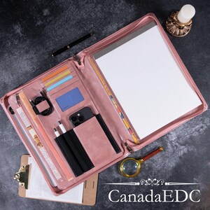 May include: A pink leather portfolio with multiple compartments, including card slots, a phone holder, and a notepad. A black pen, charging cables, and a magnifying glass are also visible. The text "CanadaEDC" is at the bottom.