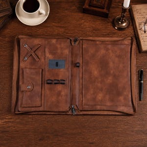 May include: A brown leather portfolio organizer featuring a zippered closure, an interior pocket with a snap button, three elastic pen loops, a card slot, and a cross-strap for securing items. The interior is lined with matching brown material.