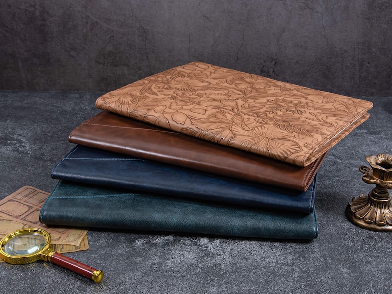 May include: A stack of four rectangular folders or portfolios. The top one is light tan with an embossed floral pattern. The three underneath are solid, smooth-textured in shades of dark brown, navy blue, and dark teal, each with stitched edges.