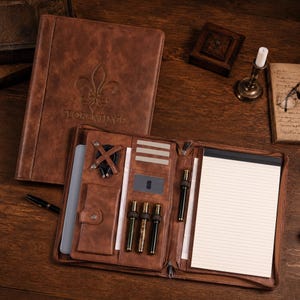 May include: A brown leather portfolio with a debossed fleur-de-lis and the text FLORENTINCE on the cover. The interior features a lined notepad, slots for pens, a small pocket with a snap closure, card slots, and a cable organizer strap.