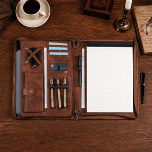 May include: A brown leather zippered portfolio organizer featuring a lined notepad, three pen loops, a card slot, a small snap-button pocket, and a cable organizer. The interior includes multiple slots for cards and documents, with a black pen attached.