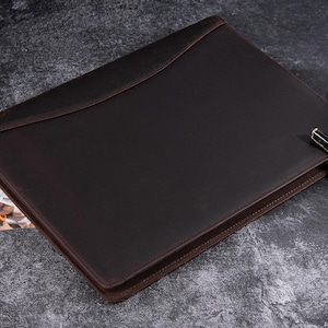 May include: A dark brown leather portfolio with a curved flap closure. The portfolio has a stitched border and a pen resting on top. A newspaper is partially visible in the background.