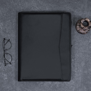 Könnte beinhalten: Ein schwarzes Lederportfolio mit Klappenverschluss und einer Reißverschlusstasche. Das Portfolio liegt flach auf einer grauen Oberfläche.