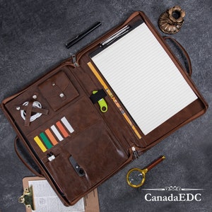 May include: A brown leather portfolio with a zipper closure, a notepad, and multiple pockets for pens, a phone, and other accessories. The portfolio has a handle and a clip for holding papers.