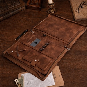 May include: A brown leather document portfolio featuring an open interior with a zippered closure, a clear ID window, three elastic pen loops, a cross-strap for securing items, and a snap-button pocket for organizing documents and stationery.