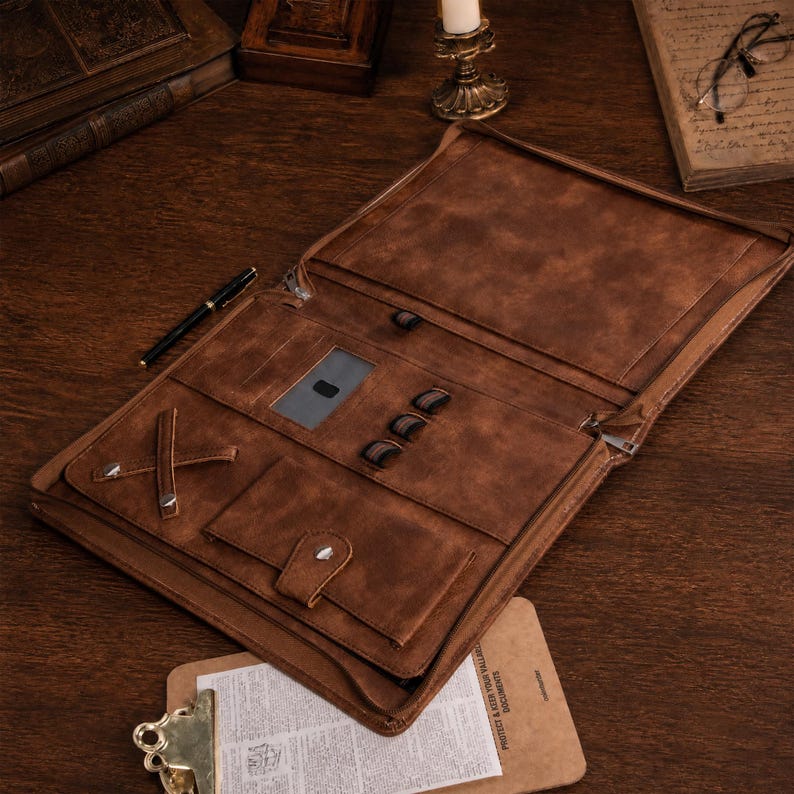 May include: A brown leather document portfolio featuring an open interior with a zippered closure, a clear ID window, three elastic pen loops, a cross-strap for securing items, and a snap-button pocket for organizing documents and stationery.