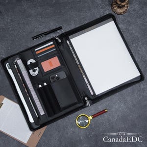 Könnte beinhalten: Schwarzes Leder-Zipper-Portfolio mit mehreren Taschen und Steckplätzen für die Organisation. Das Portfolio enthält einen Notizblock, einen Stift, einen Telefonhalter und anderes Zubehör.