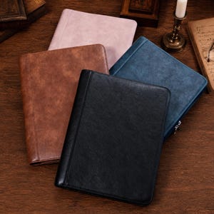 May include: Four rectangular portfolios with a textured faux leather finish in black, brown, blue, and light pink. Each portfolio features a vertical stitched seam along the left side and a smooth, solid-colored front cover with a subtle grain pattern.