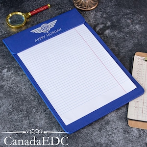 May include: A blue leather-like clipboard featuring a top flap with the white printed text AVERY MORGAN and a winged globe logo. The clipboard holds a white legal pad with blue horizontal lines and a single red vertical margin line on the right side.