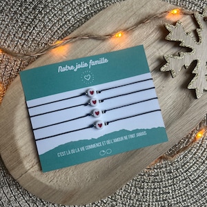 Puede incluir: Un conjunto de cinco pulseras de amistad con cuentas blancas y corazones rojos. Las pulseras están empaquetadas en una tarjeta con el texto "Notre jolie famille" y "C'EST LÀ OÙ LA VIE COMMENCE ET OÙ L'AMOUR NE FINIT JAMAIS".