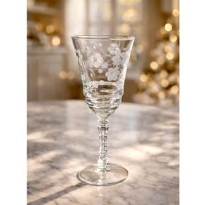 Puede incluir: Una copa de cristal transparente grabada con un diseño floral. La copa tiene un cuenco acampanado, un tallo detallado y una base redonda. El diseño grabado presenta delicadas flores y hojas. El vaso está sobre una superficie de mármol.