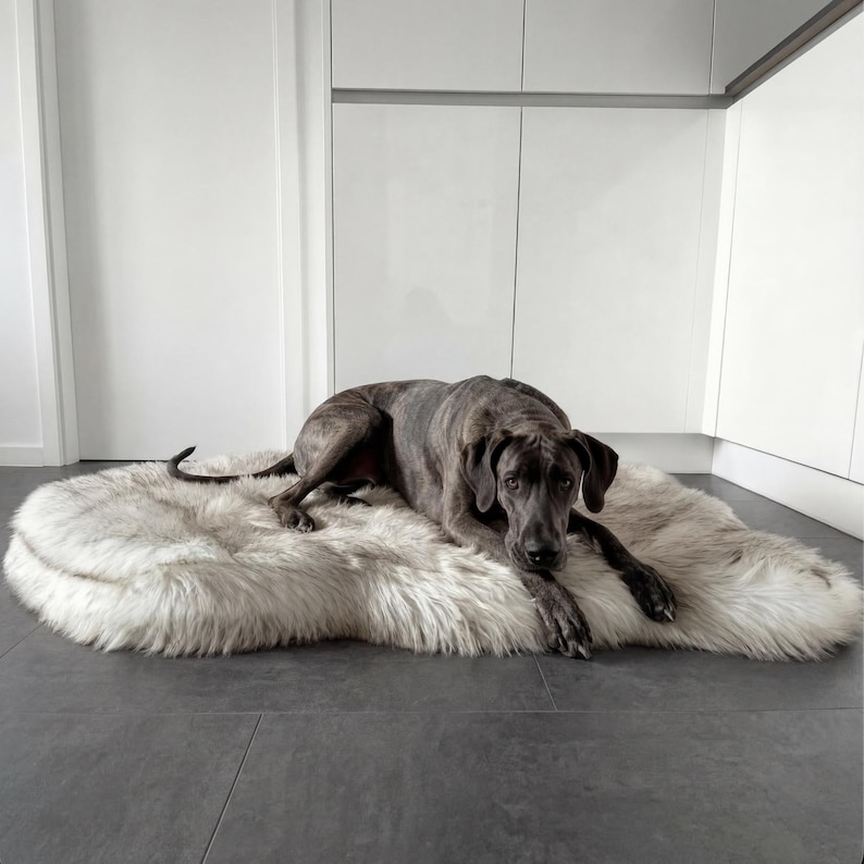 K&ouml;nnte beinhalten: Ein gro&szlig;er gestromter Hund liegt auf einem flauschigen, cremefarbenen Kunstfell-Haustierbett. Das Fell des Hundes hat ein Muster aus dunklen Streifen auf grauem Grund. Das Bett ist oval und liegt auf einem dunkelgrauen Boden, mit wei&szlig;en Schr&auml;nken im Hintergrund.