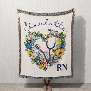 Puede incluir: Una manta tejida con fondo blanco y un diseño de corazón floral. El corazón está formado por flores coloridas y un estetoscopio. El texto "Charlotte RN" está impreso en azul en la parte inferior de la manta.